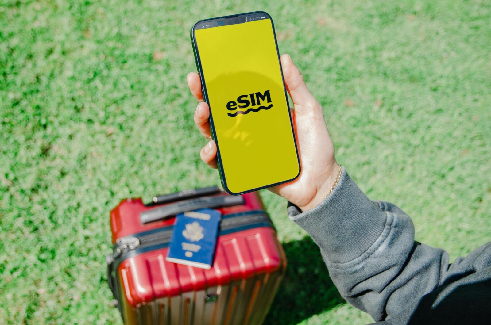 Traveler in Los Angeles holding a smartphone with eSIM near a red suitcase on grass.