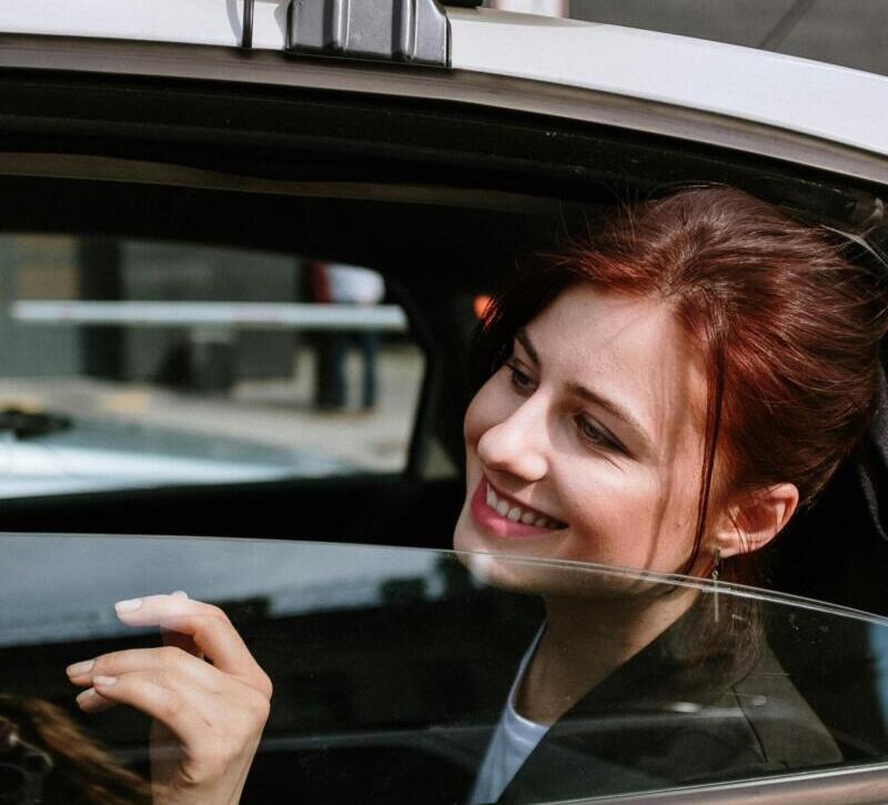 A cheerful woman smiles from the backseat of a moving taxi, city street view.