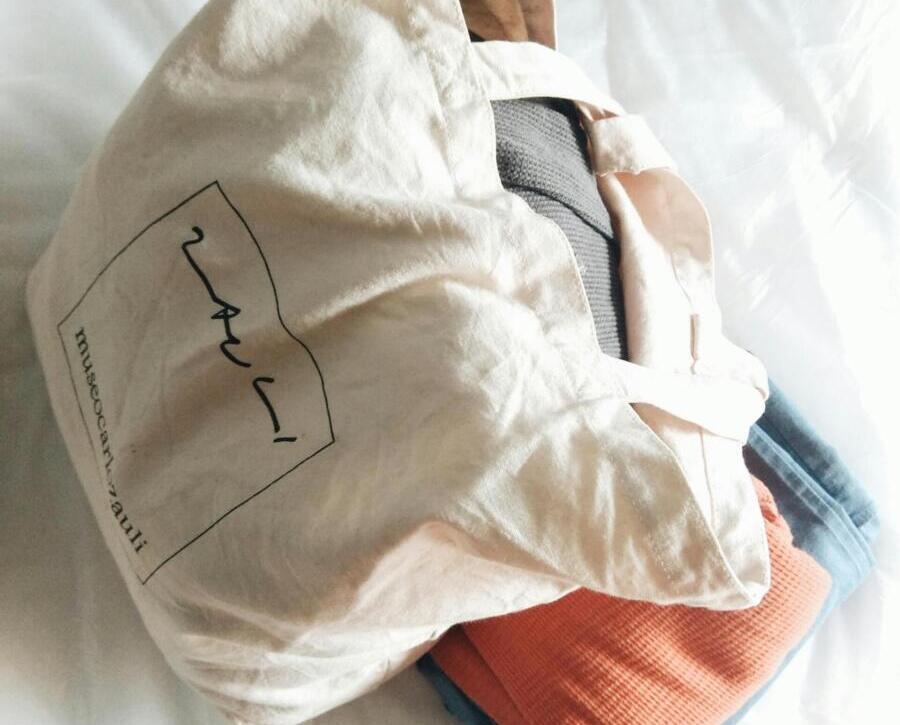 A natural cotton tote bag filled with clothes resting on a neatly made bed indoors.