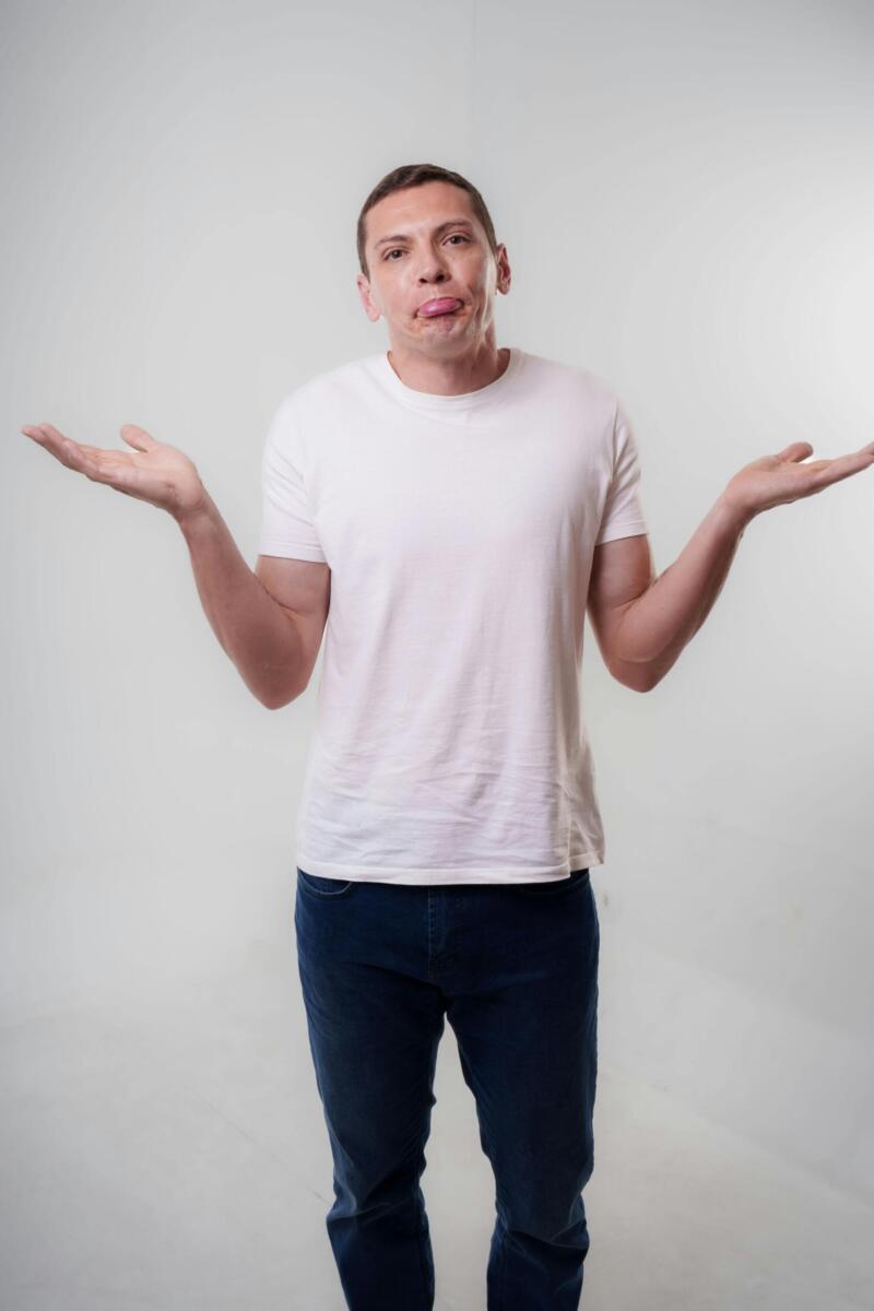 A young adult man in casual clothing shrugs with a neutral expression in a studio setting.