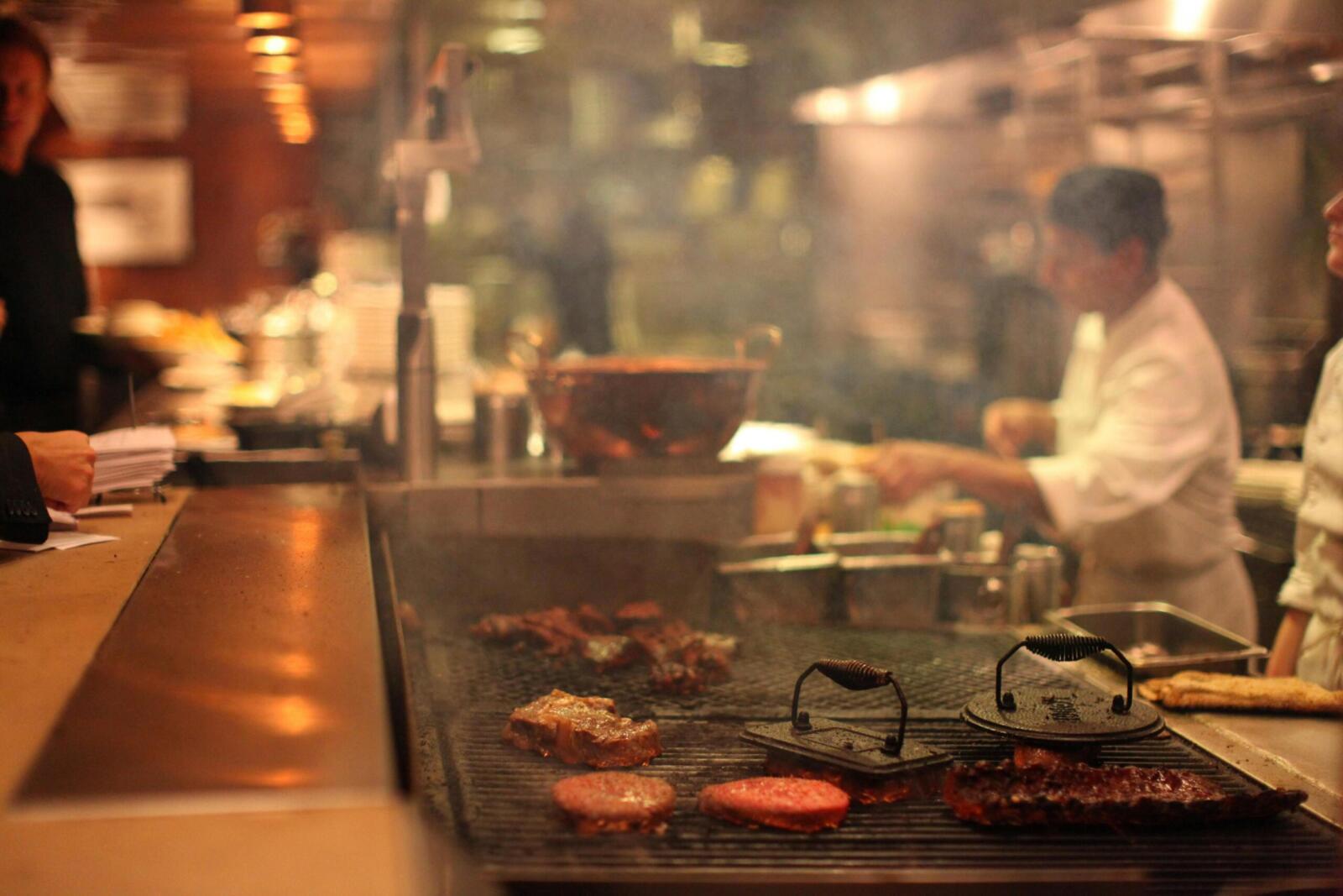Chefs grilling various meats in a bustling restaurant kitchen, focused on service.