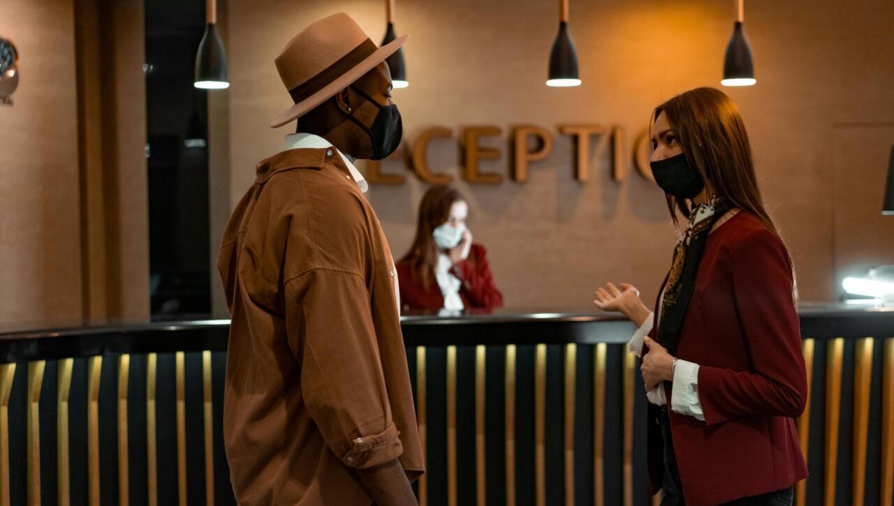 Two people in face masks interacting at a hotel reception desk during the COVID-19 pandemic.
