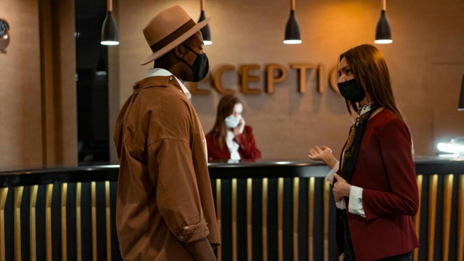 Two people in face masks interacting at a hotel reception desk during the COVID-19 pandemic.