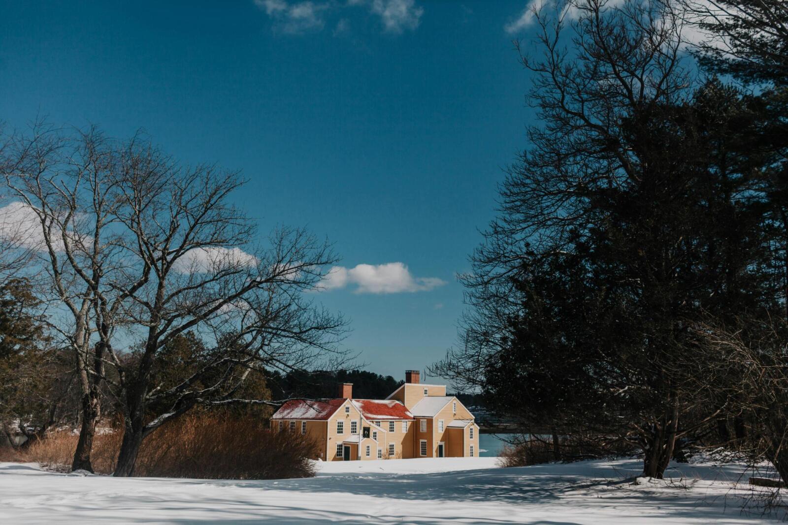 Winter scene with a quaint yellow house surrounded by snow and bare trees in New Hampshire.