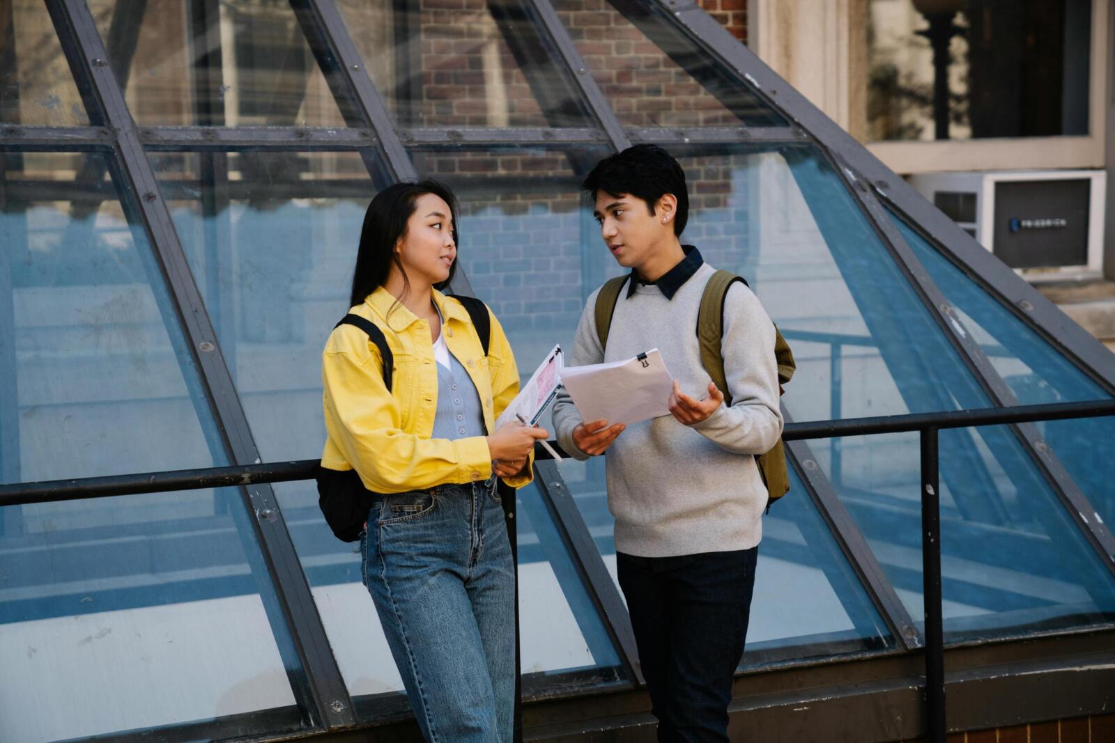 Two young students conversing outdoors with papers and backpacks, suggesting a college campus setting.