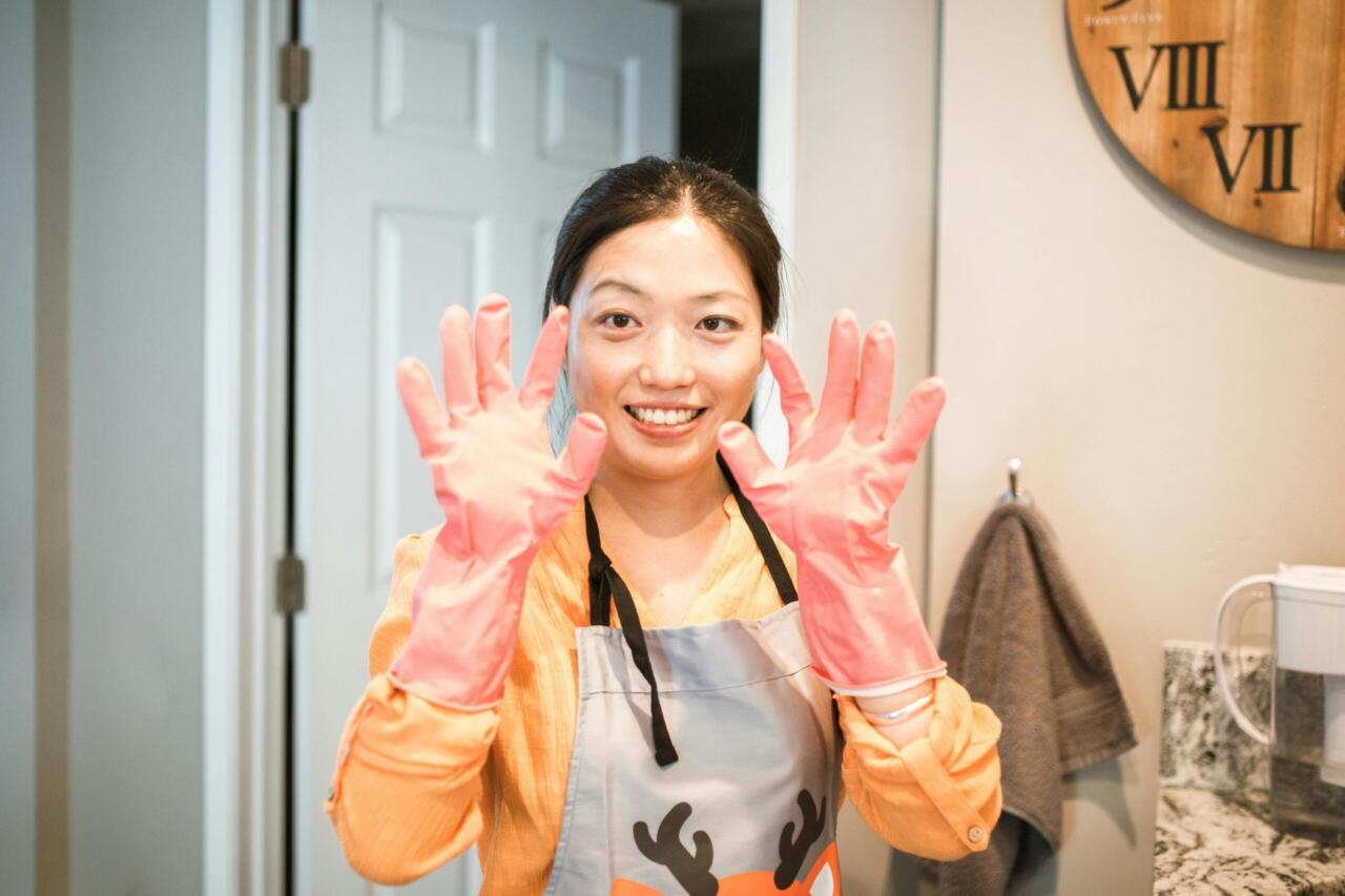 Smiling woman in kitchen apron shows pink gloves, ready for cleaning tasks.