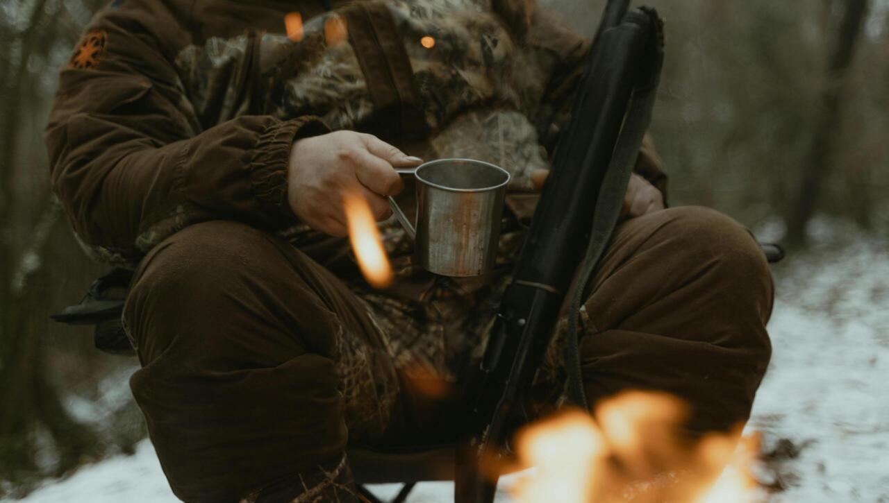 A hunter wearing camouflage sits by a campfire holding a metal mug in a snowy forest.